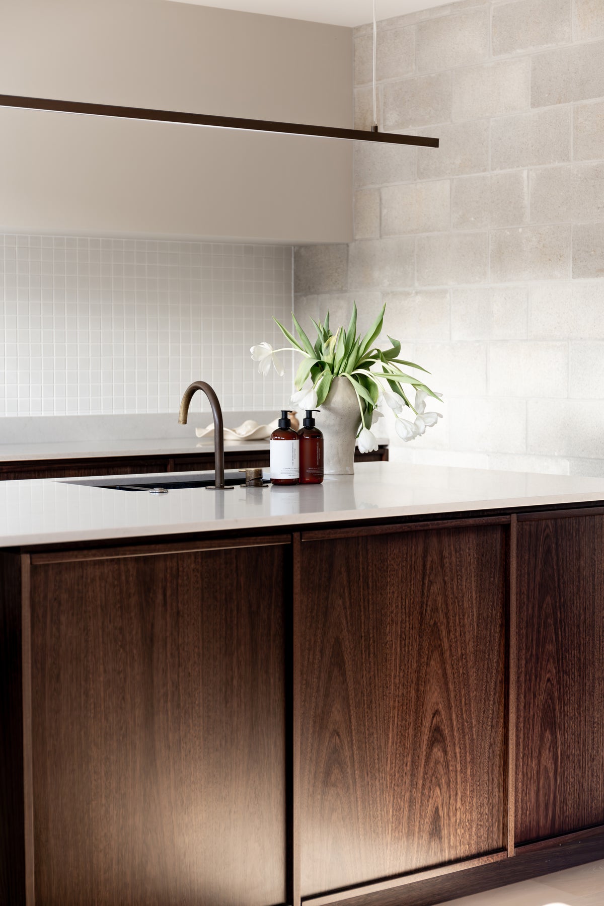 Kitchen sink set into a dark timber island, amber pump bottles and tulips on the bench