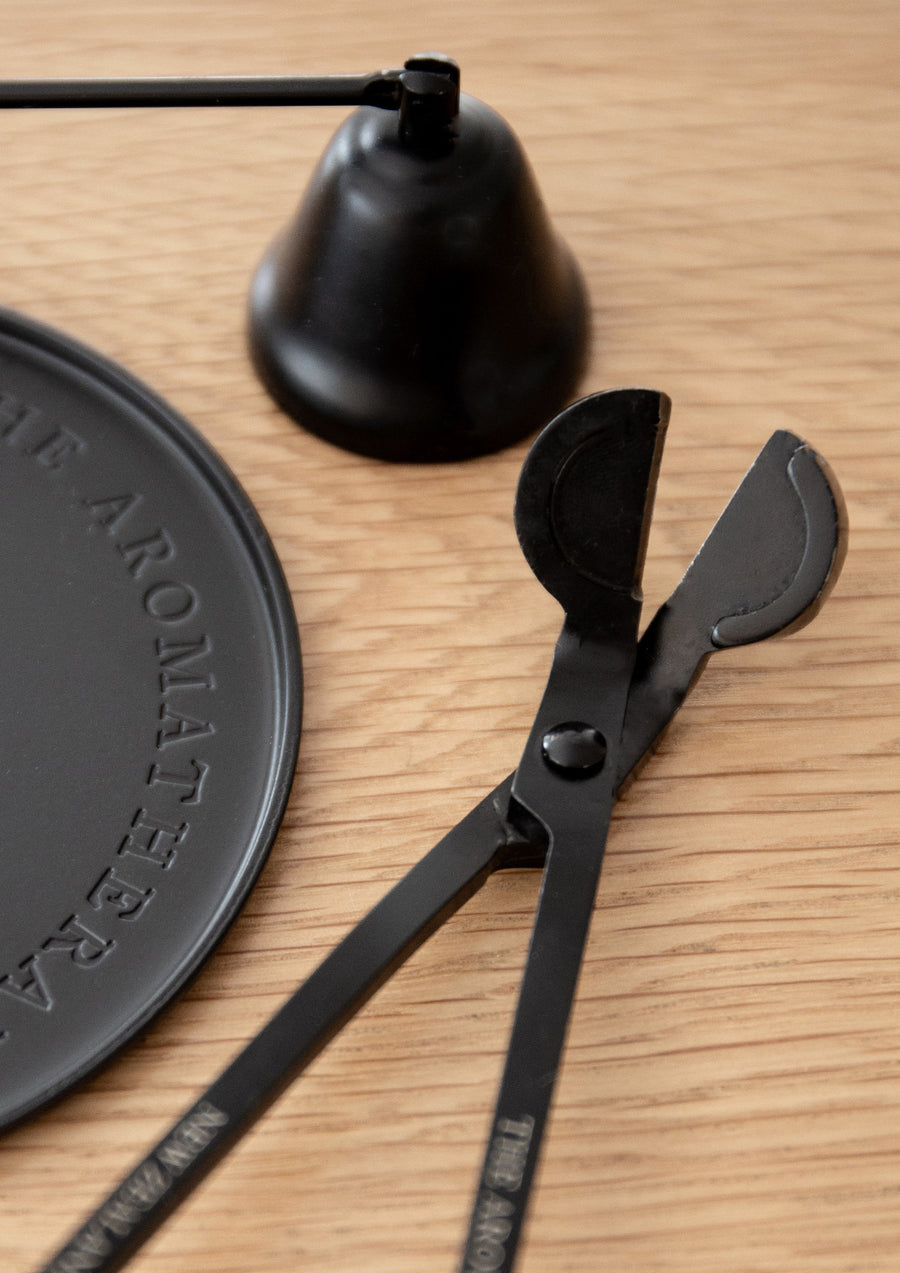 Black wick trimmer open on light oak beside a black candle snuffer and an embossed black plate