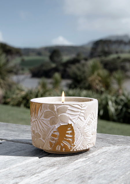 Lit leaf-embossed concrete candle on a weathered timber table with bush and a distant peak behind