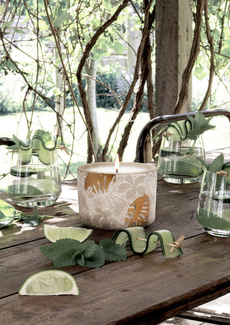 Lit outdoor candle on a rustic timber table under a vine pergola with glasses, lime wedges and mint