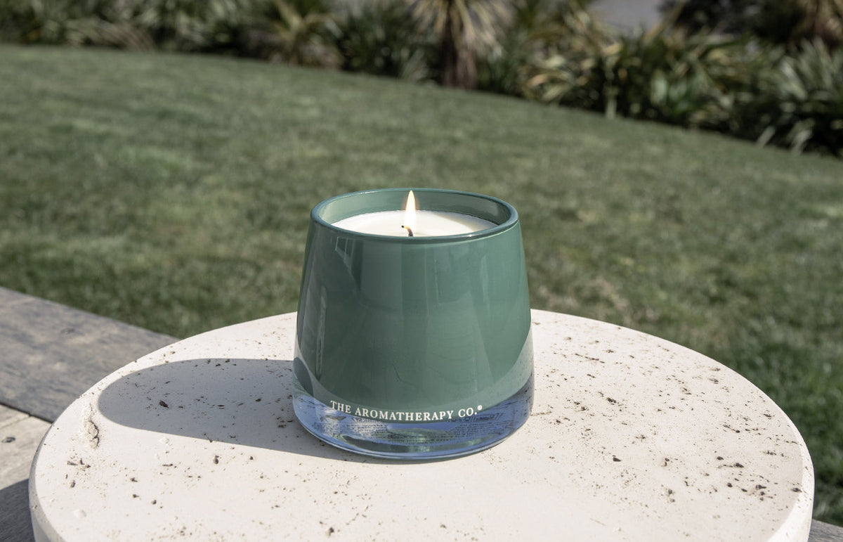 Lit green glass candle on a round stone table outdoors with a lawn and shrubs behind