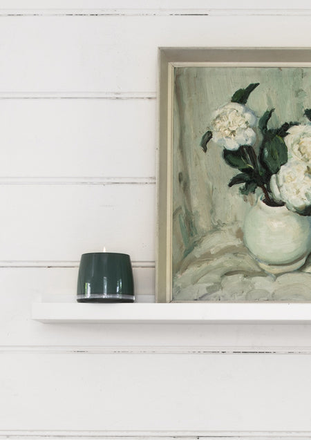 Small green candle on a white shelf below a framed floral painting