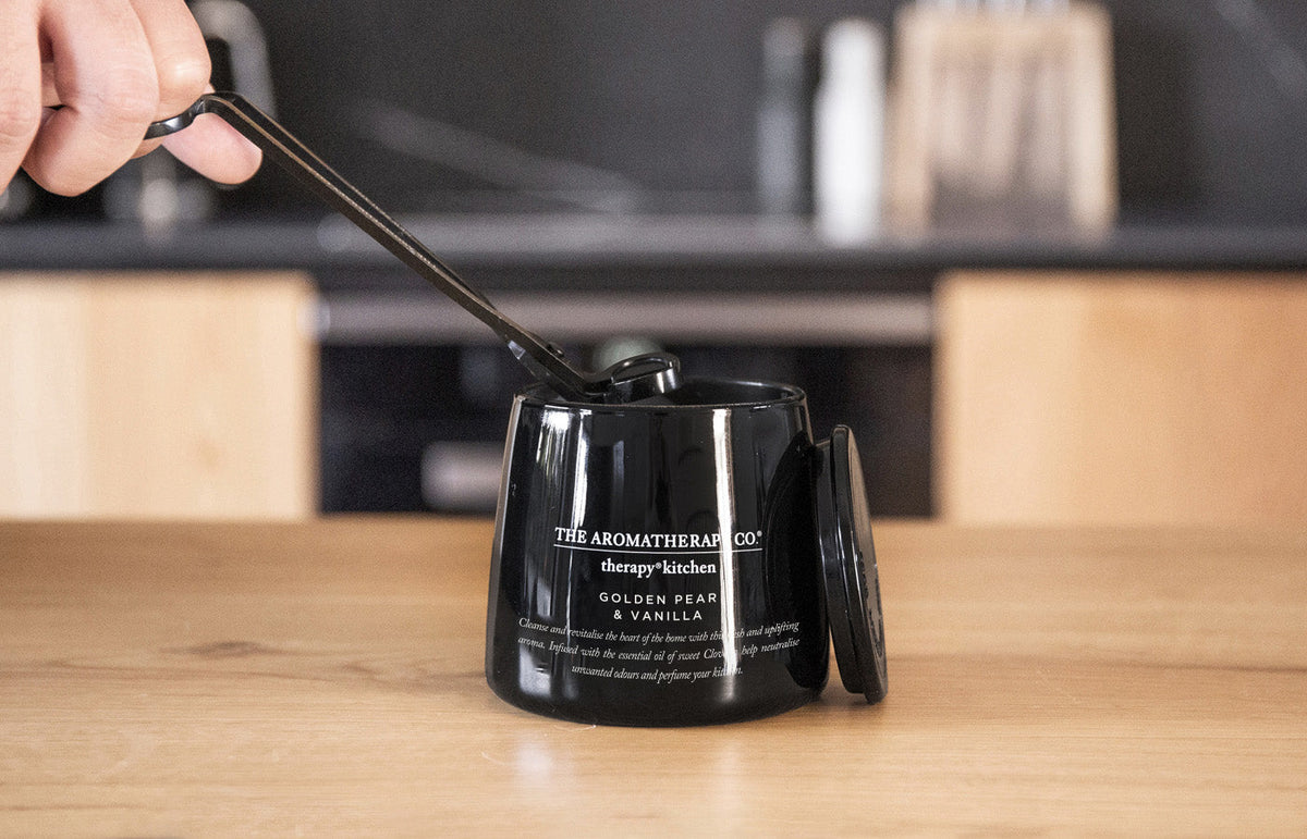 Hand lowering a snuffer over the lit Therapy Kitchen candle on a timber bench