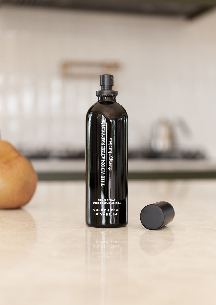 Black room spray with its cap beside it on a stone kitchen bench with a pear and cooktop behind