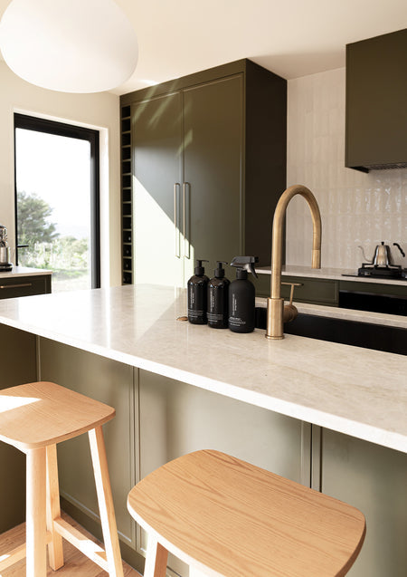 Olive green kitchen with a stone island, brass tap and three black Therapy Kitchen bottles beside timber bar stools
