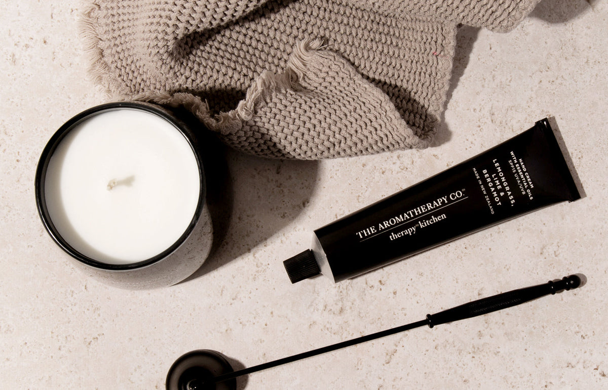 Black hand cream tube beside a candle, a wick trimmer and a knit cloth on pale stone, from above