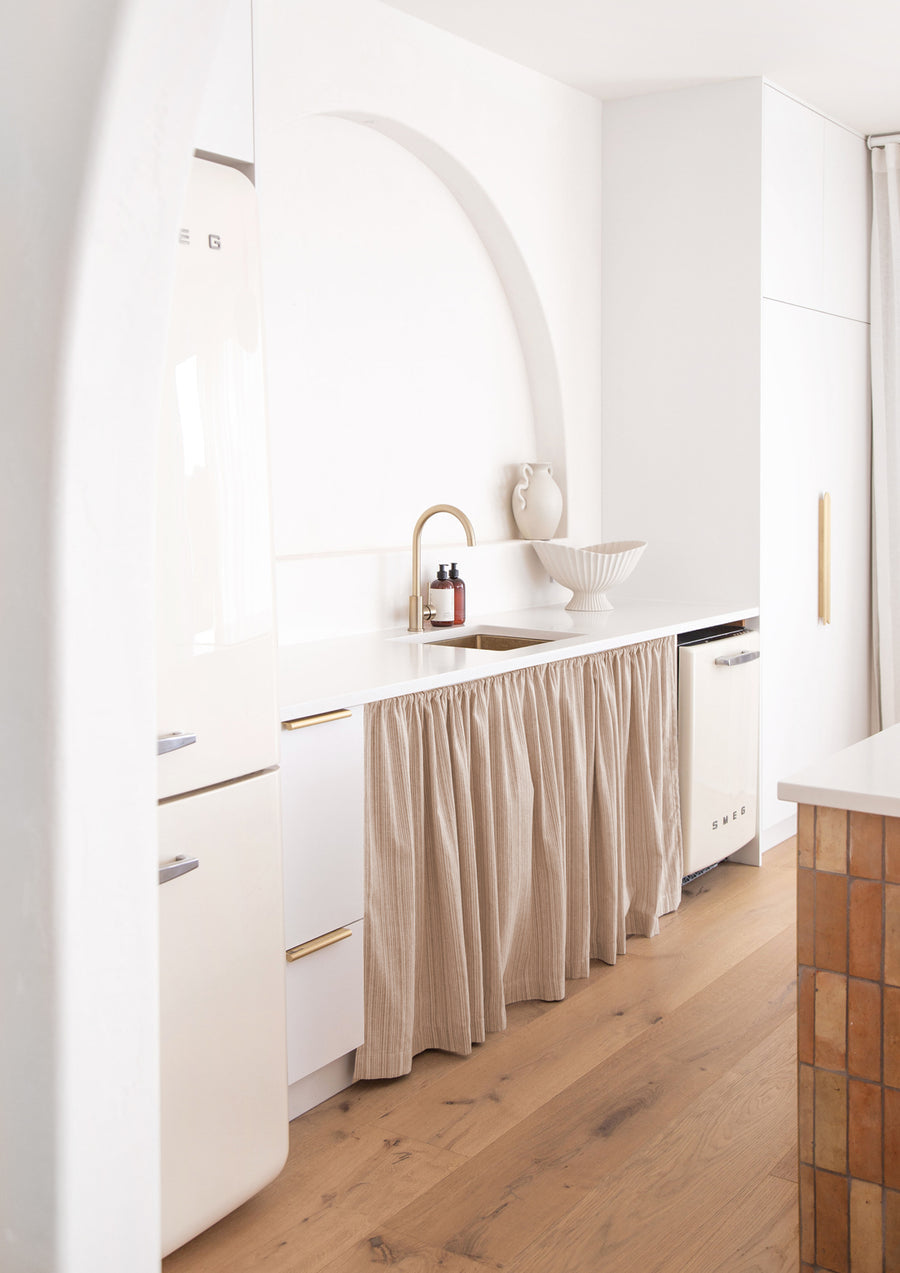 Bright white kitchen with an arched alcove, curtained under-bench storage and two small bottles by the tap