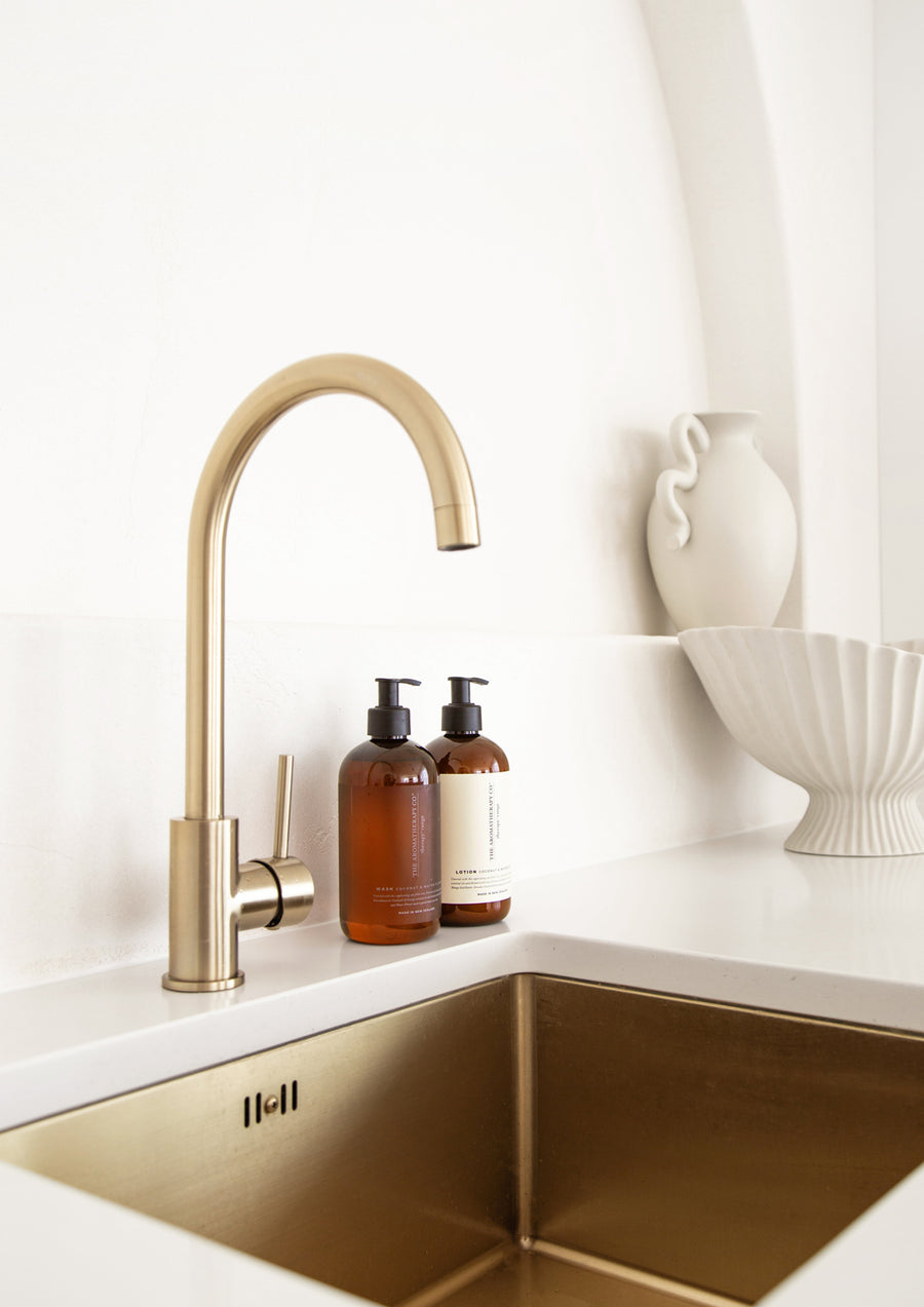 Two amber Therapy pump bottles beside a brass gooseneck tap and brass sink with white ceramics