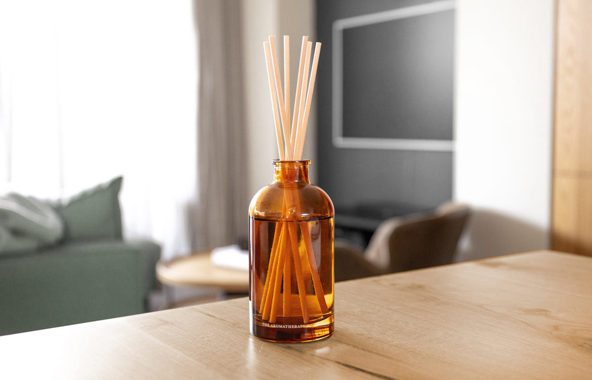 Amber reed diffuser on a timber dining table with a living room and sofa blurred behind