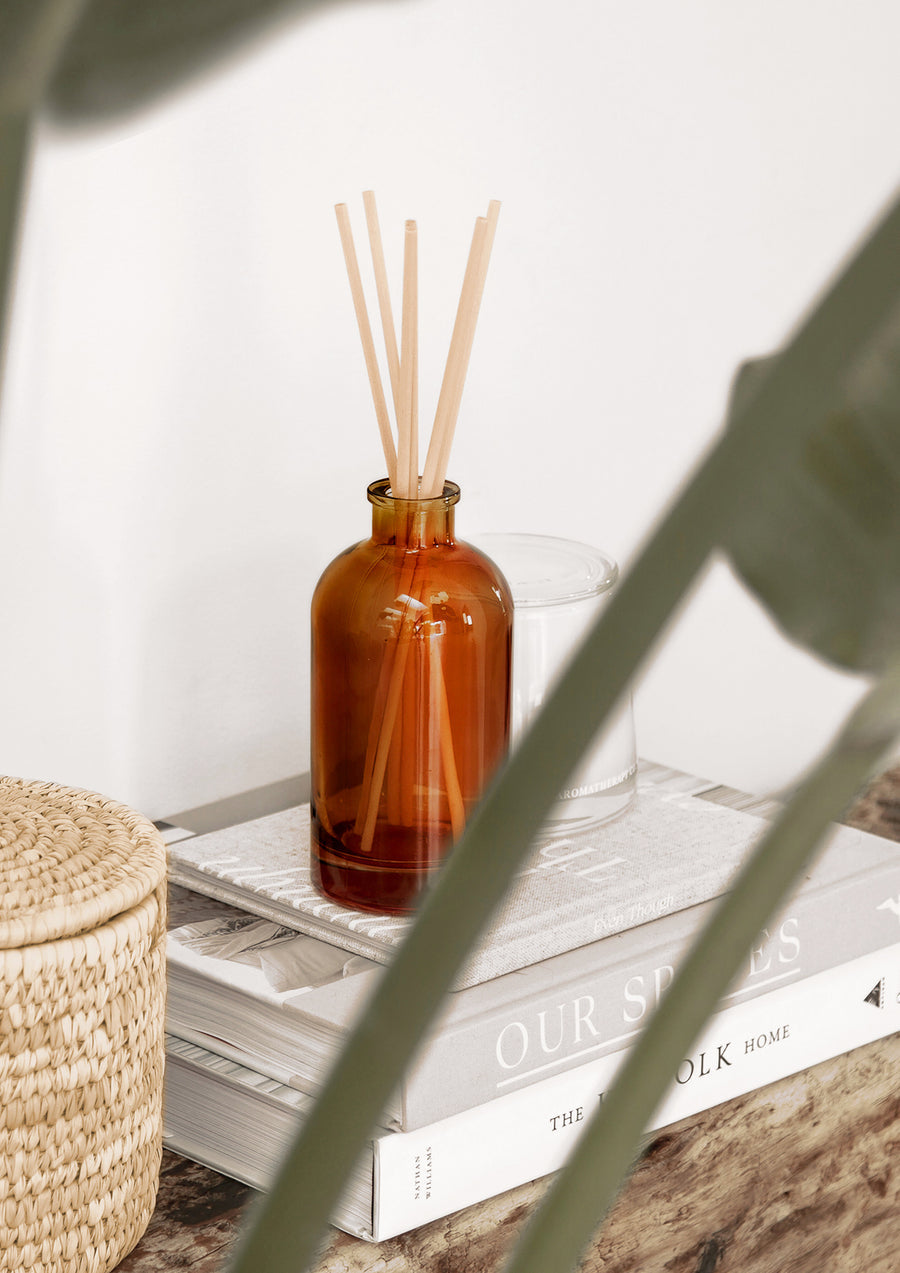 Amber reed diffuser on a stack of books beside a woven basket with a leafy stem in front