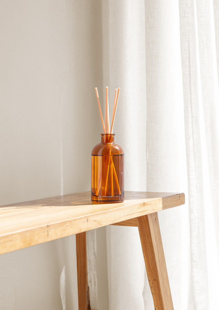 Amber reed diffuser on a light timber console against sheer white curtains