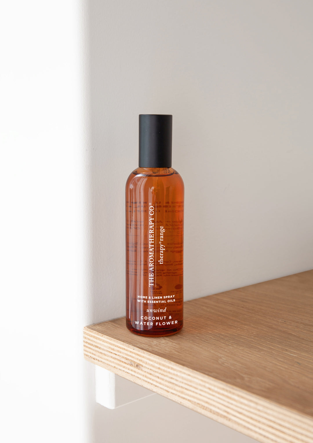 Amber room spray at the corner of a pale oak shelf against a white wall