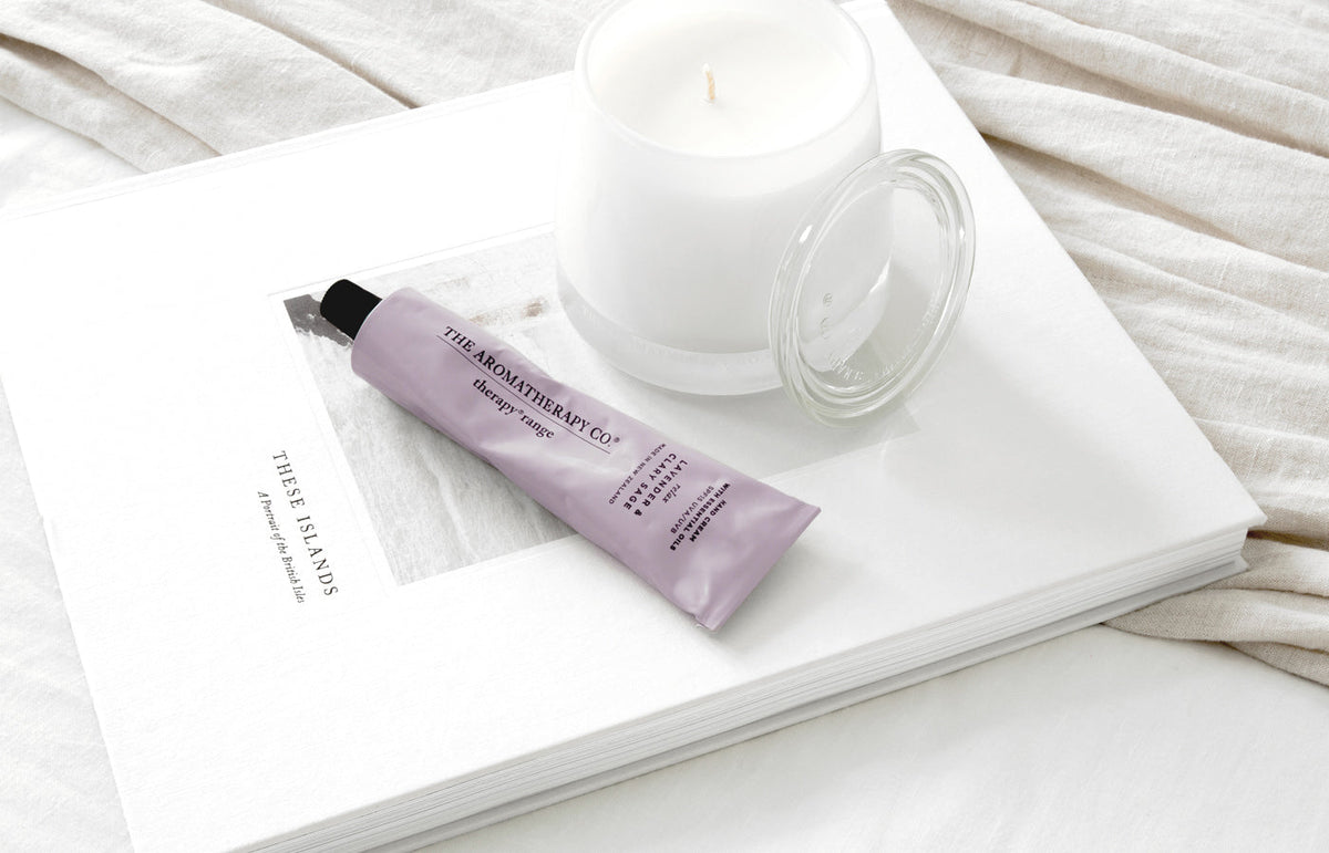 Lilac hand cream tube on an open book beside a white glass candle on linen, landscape view