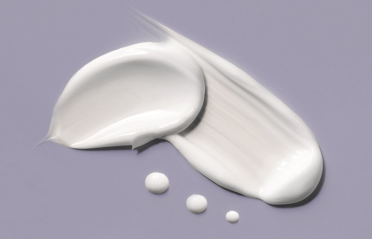 Swatch of white hand cream with three small drops on a lilac grey background