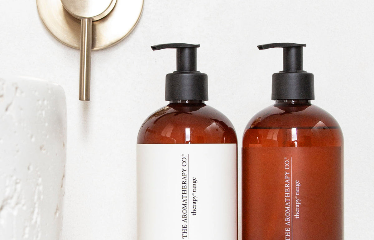Close crop of the Therapy lotion and wash bottles below a brushed brass tap, landscape view