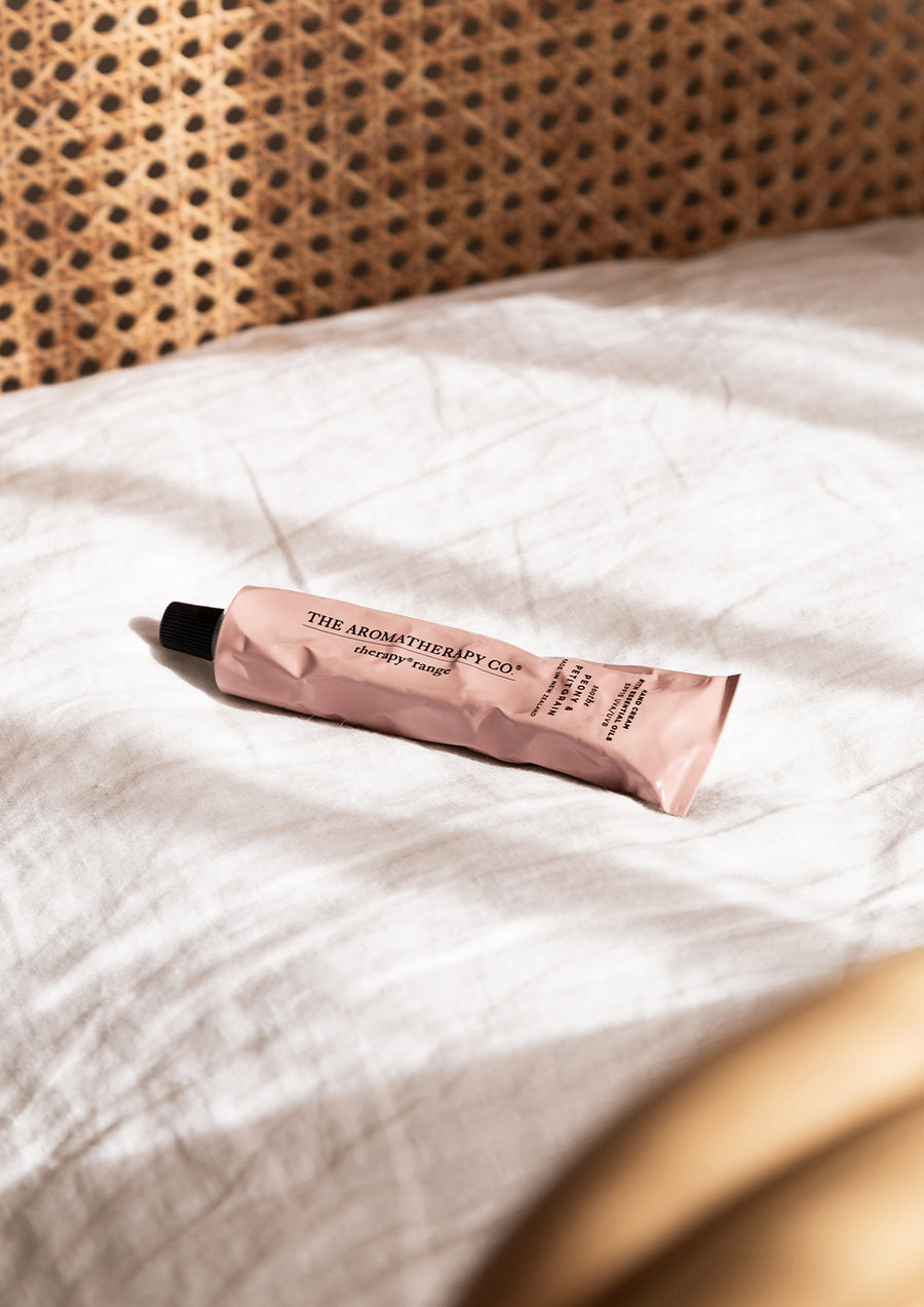 Blush hand cream tube lying on white linen beside a rattan headboard in sunlight