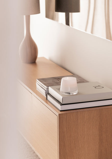 White glass candle on a stack of books on a pale oak sideboard below a ceramic table lamp