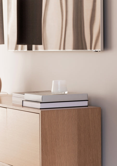 Small white candle on stacked books on an oak cabinet below a framed abstract print