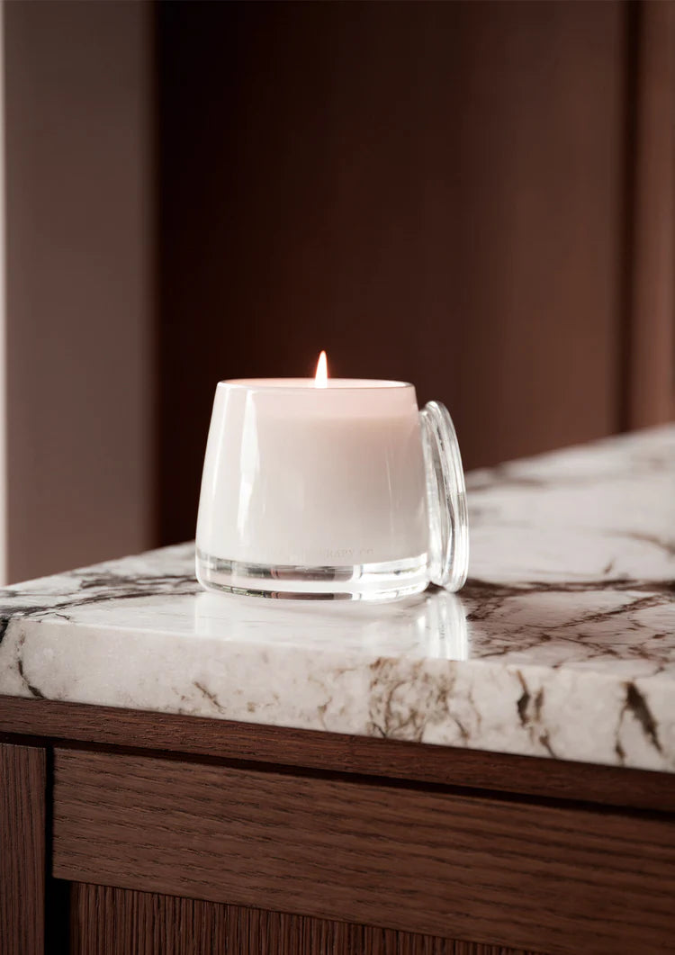Lit Embrace candle with its glass lid on a veined marble bench edge above a dark timber cabinet