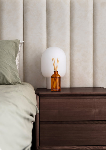Amber reed diffuser and a white mushroom lamp on a dark timber bedside table by an upholstered bedhead