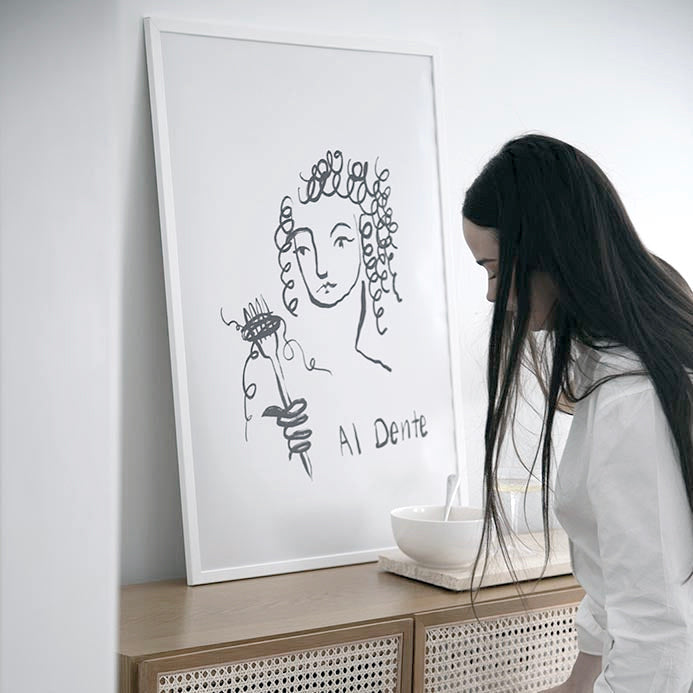 Al Dente print in a white frame on a rattan sideboard beside a woman and a pasta bowl