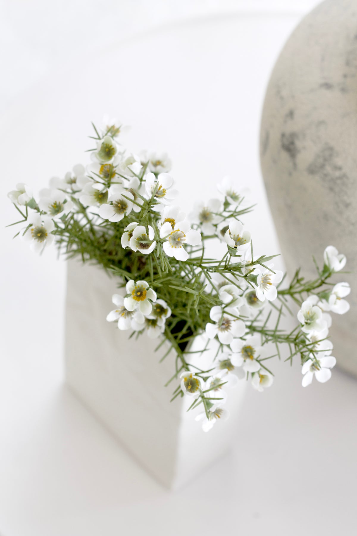 White Alba wax flower faux bush arranged in a white geometric ceramic vase beside a stone sphere on a neutral surface