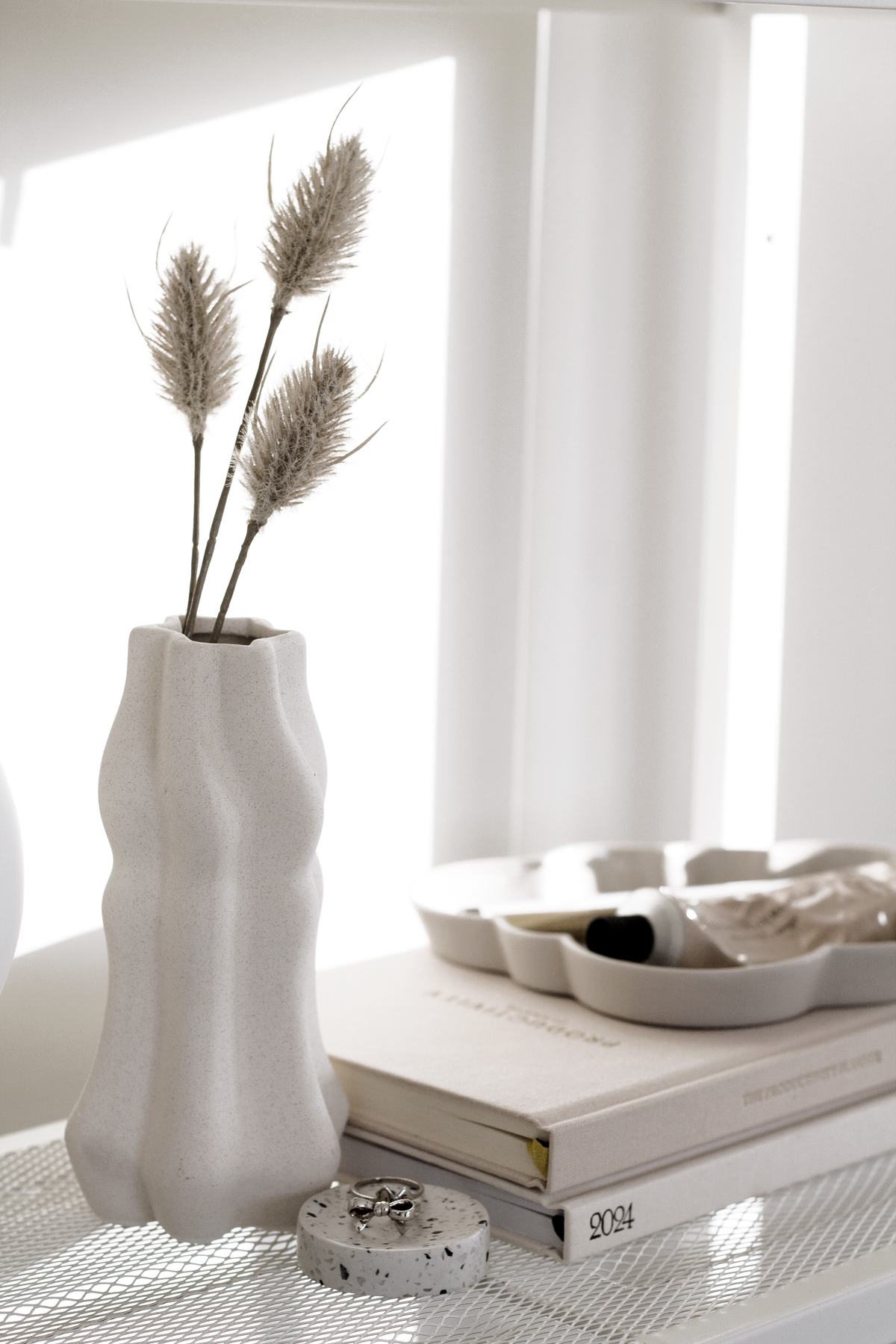 Faux bunny tails in a white wavy vase beside books and a scalloped dish on a shelf