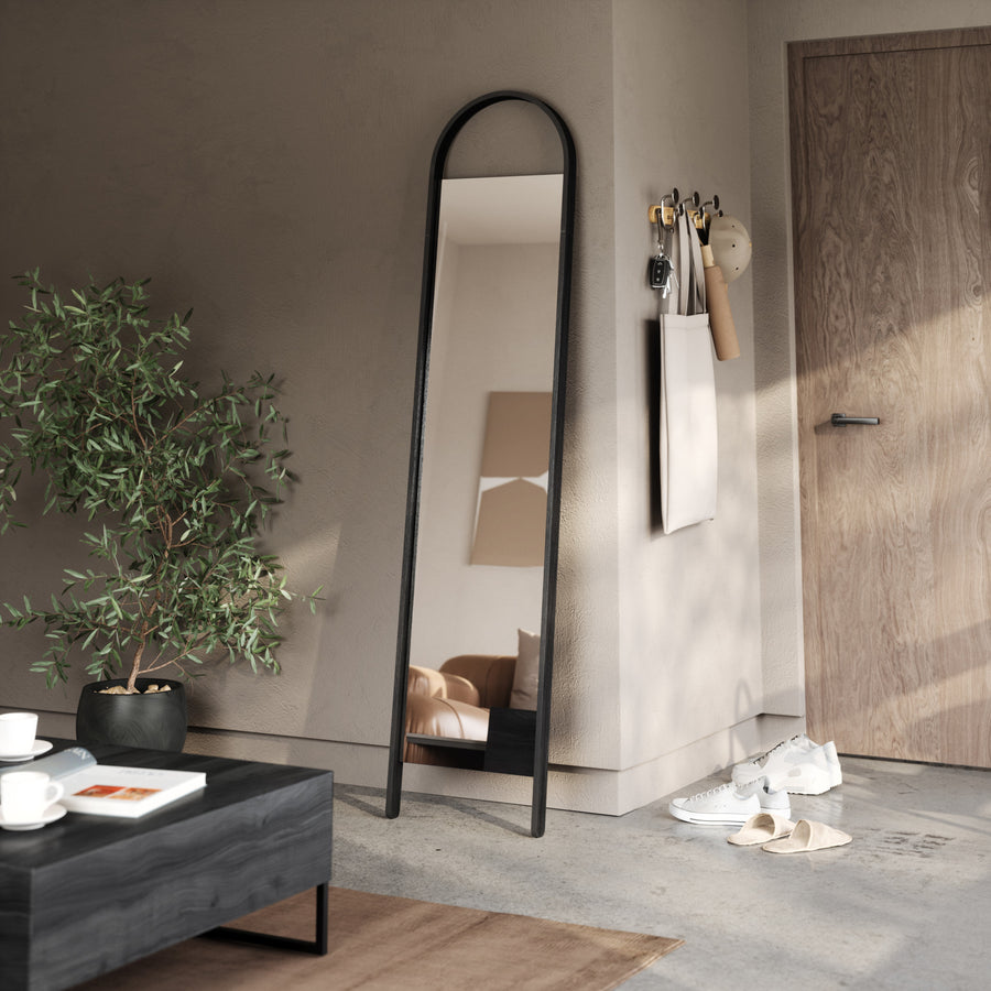 Umbra Bellwood leaning mirror in black in a bedroom corner beside a plant and bench