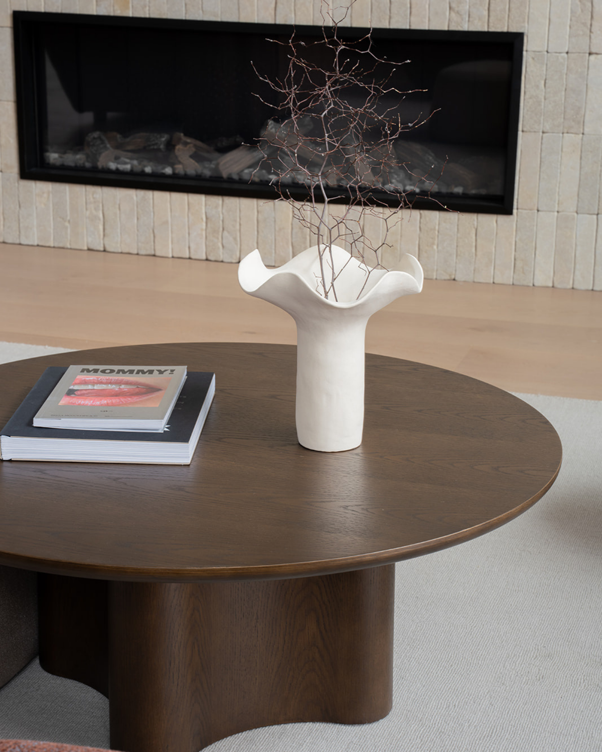 Ivory vase holding dried twigs on a round dark timber coffee table in front of a fireplace