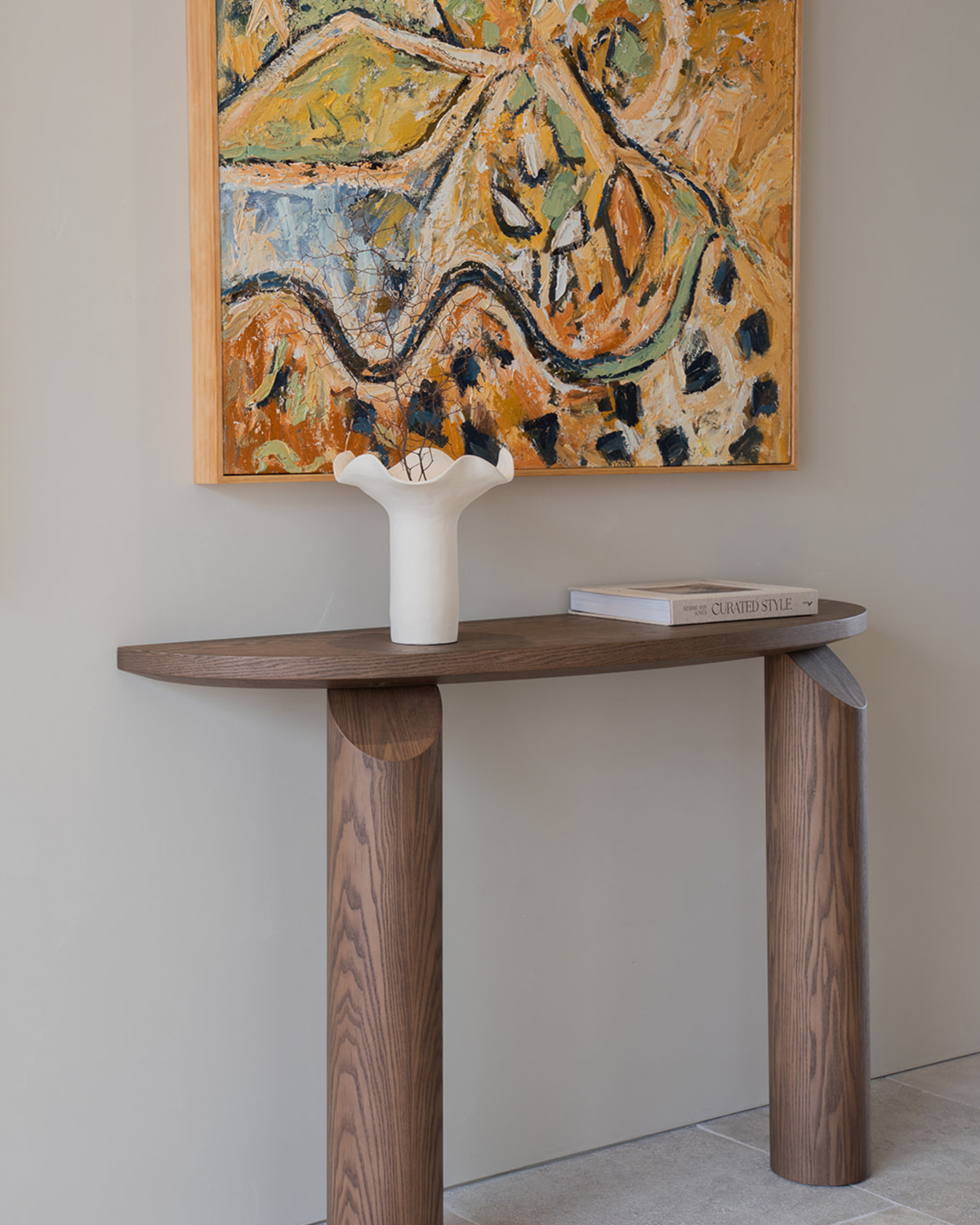Ivory vase on a curved timber console table beneath a framed abstract painting in ochre tones