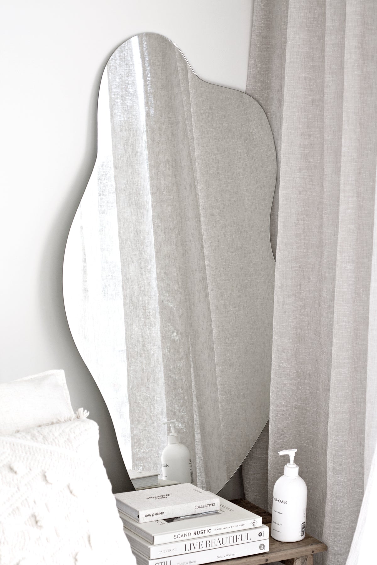 Organic blob-shaped full-length mirror leaning by soft linen curtains in a neutral, Scandi-minimal bedroom.