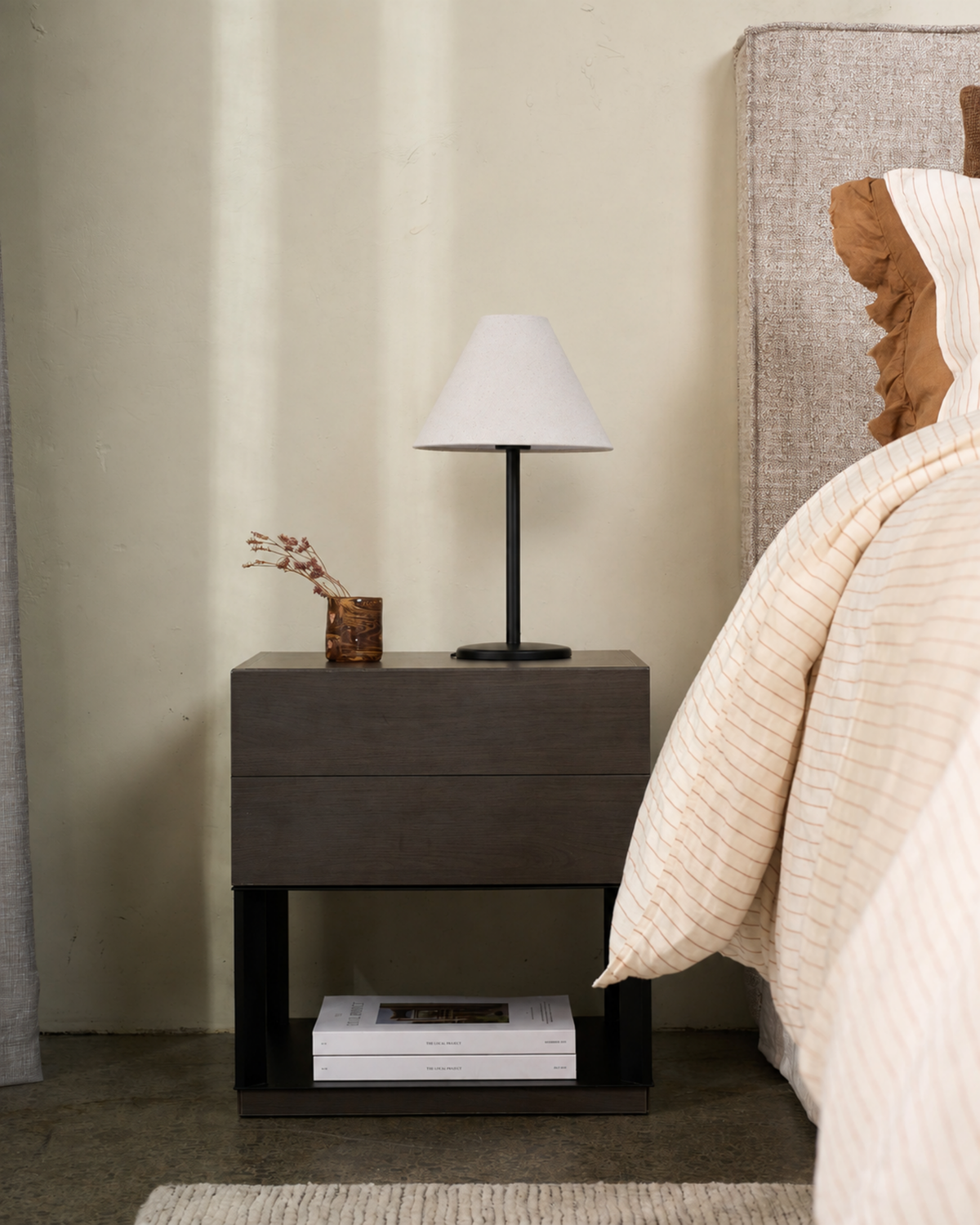 Lamp on a dark timber bedside table beside a bed with striped linen and a small vase of dried flowers