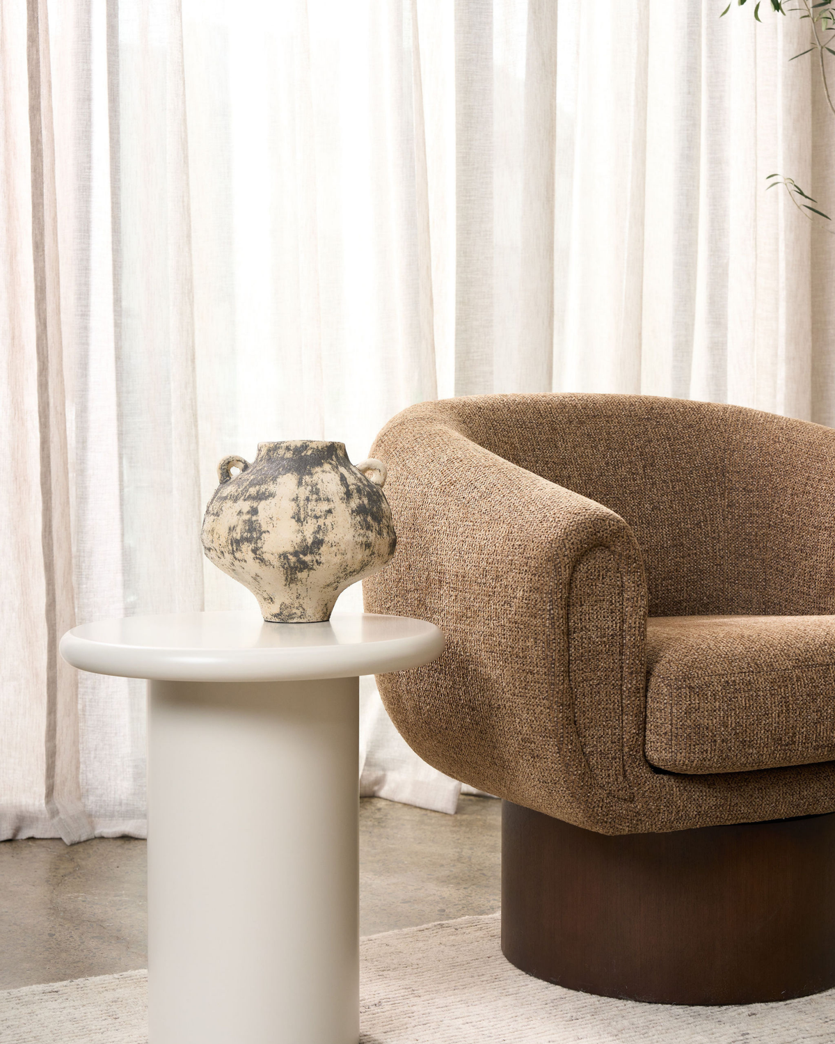 Small vase on a round cream side table beside a brown textured swivel armchair and sheer curtains