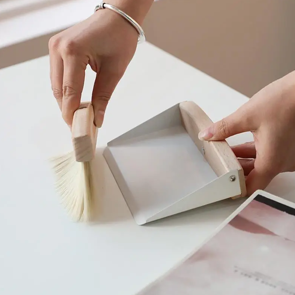 Hands sweeping crumbs from a white desk into the cream tabletop dustpan