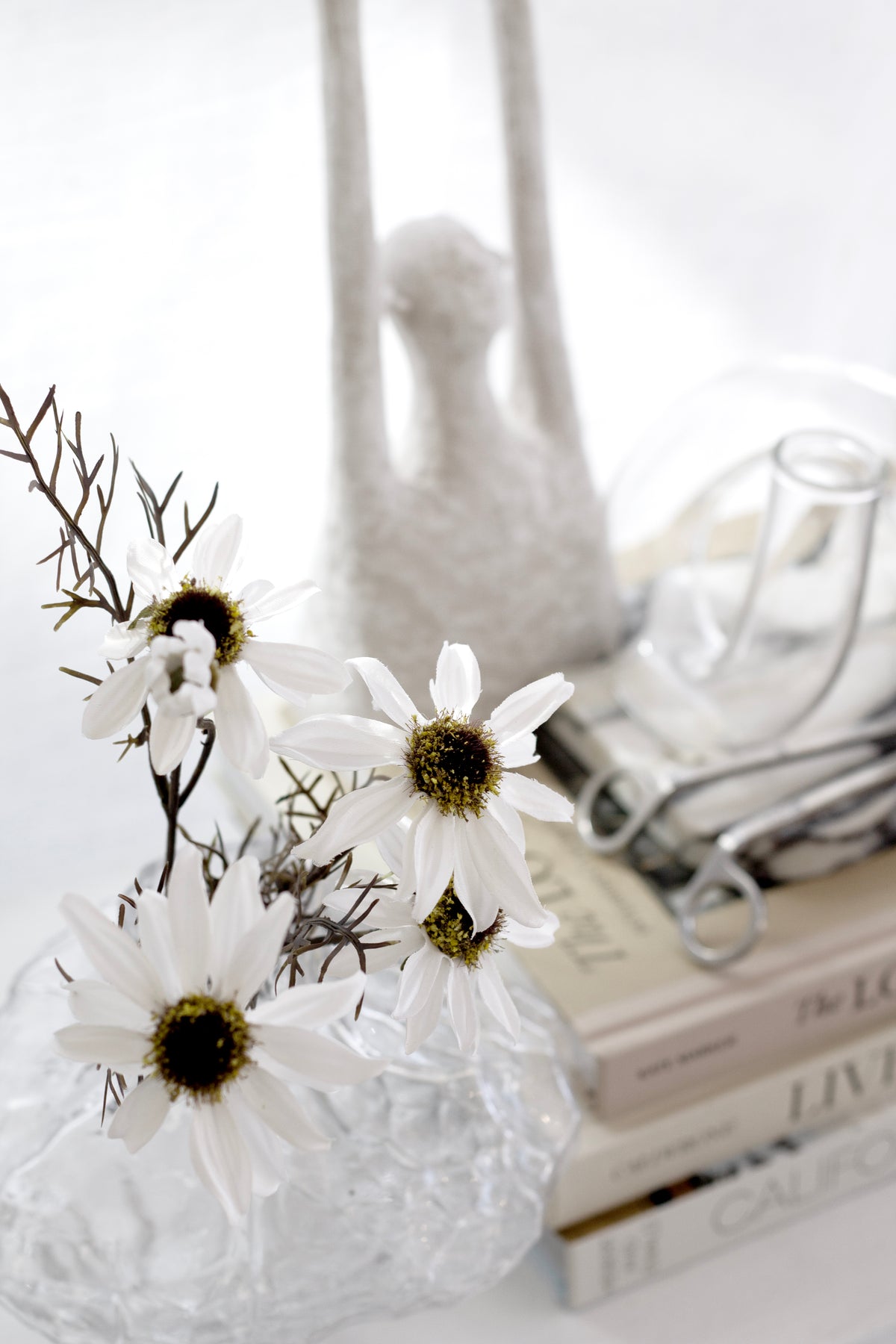 White artificial echinacea flower stems styled in a clear sculptural vase on stacked neutral books.