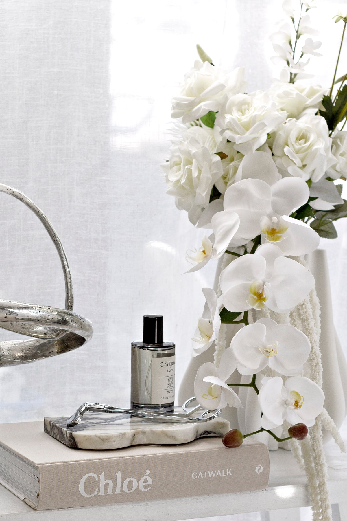 Black Ecoya Celebration Room Spray styled with white orchids, books, and a silver decorative object on a bright console table.