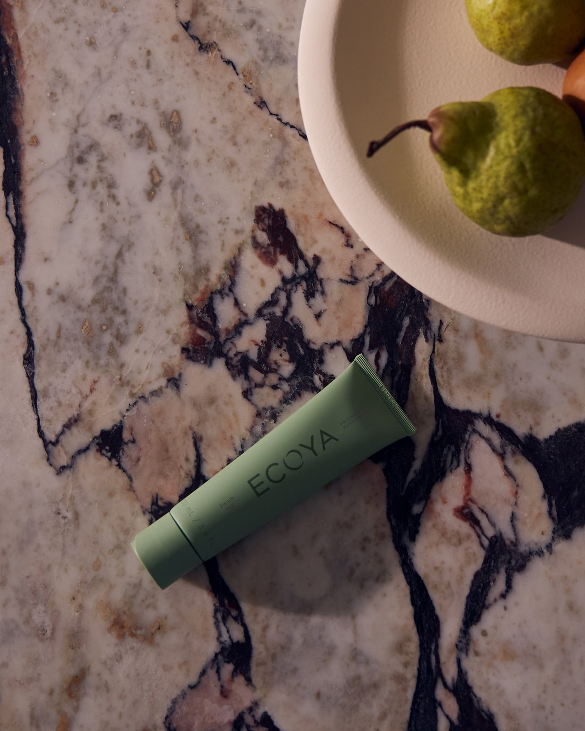 Green Ecoya hand cream tube on pink marble beside a bowl of pears