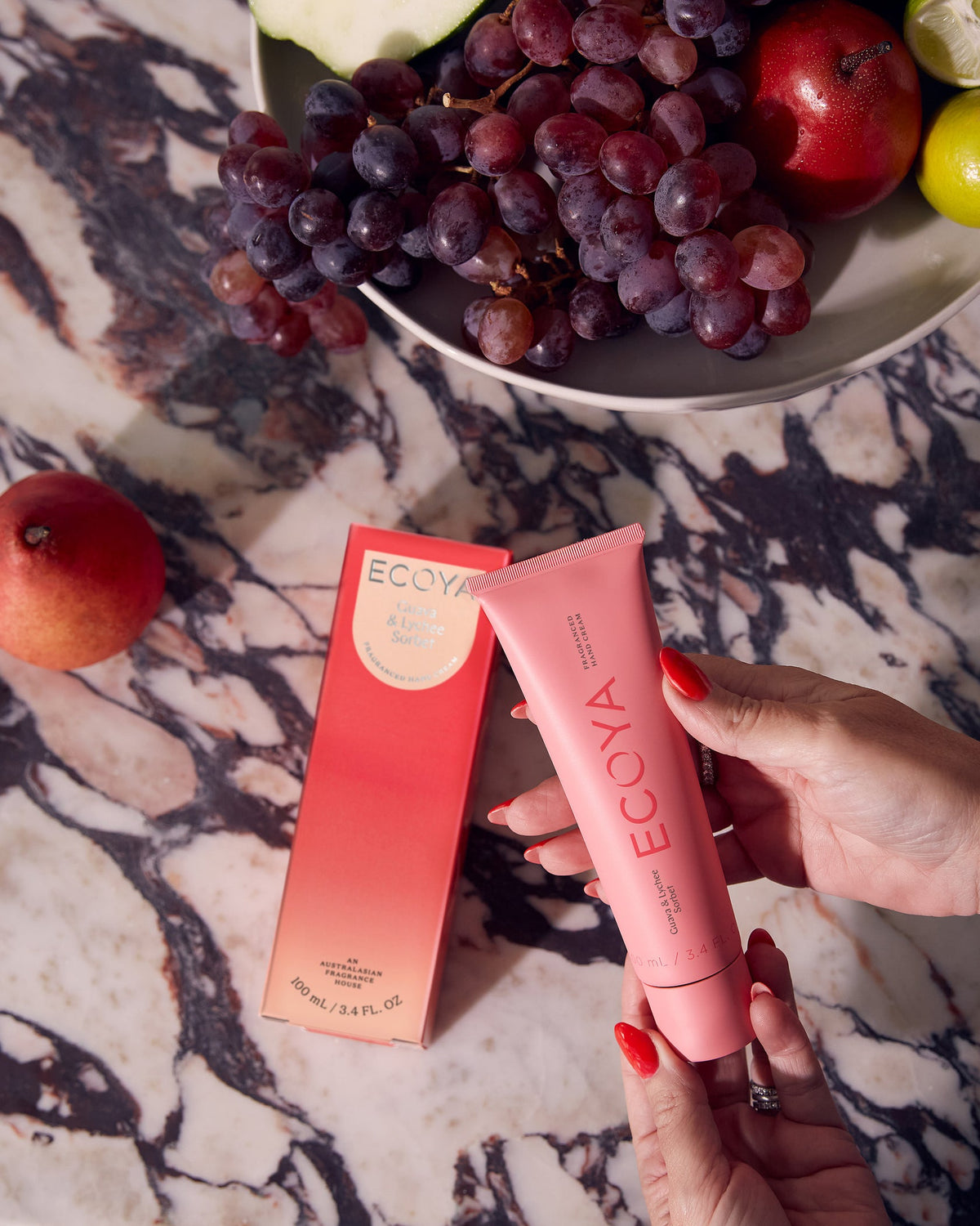 Hand holding a red Ecoya hand cream tube over a marble table with grapes and fruit