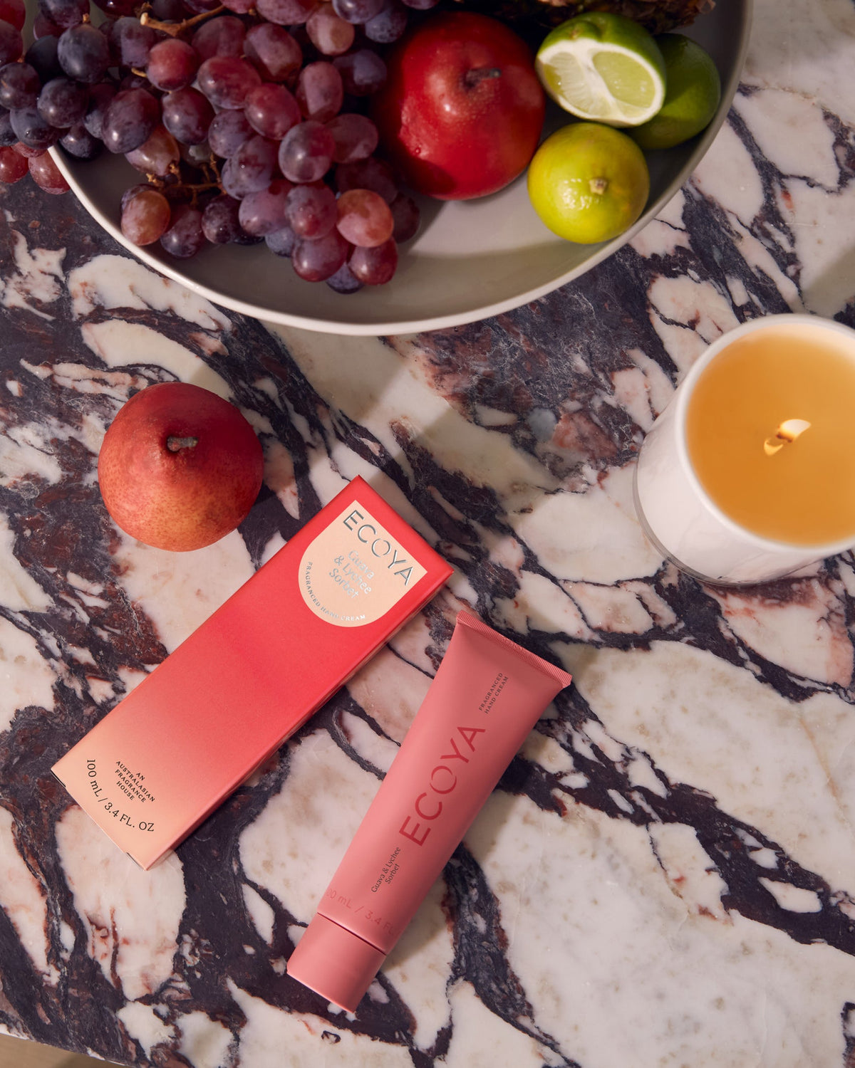 Two red Ecoya hand cream tubes on marble with grapes, apples and citrus