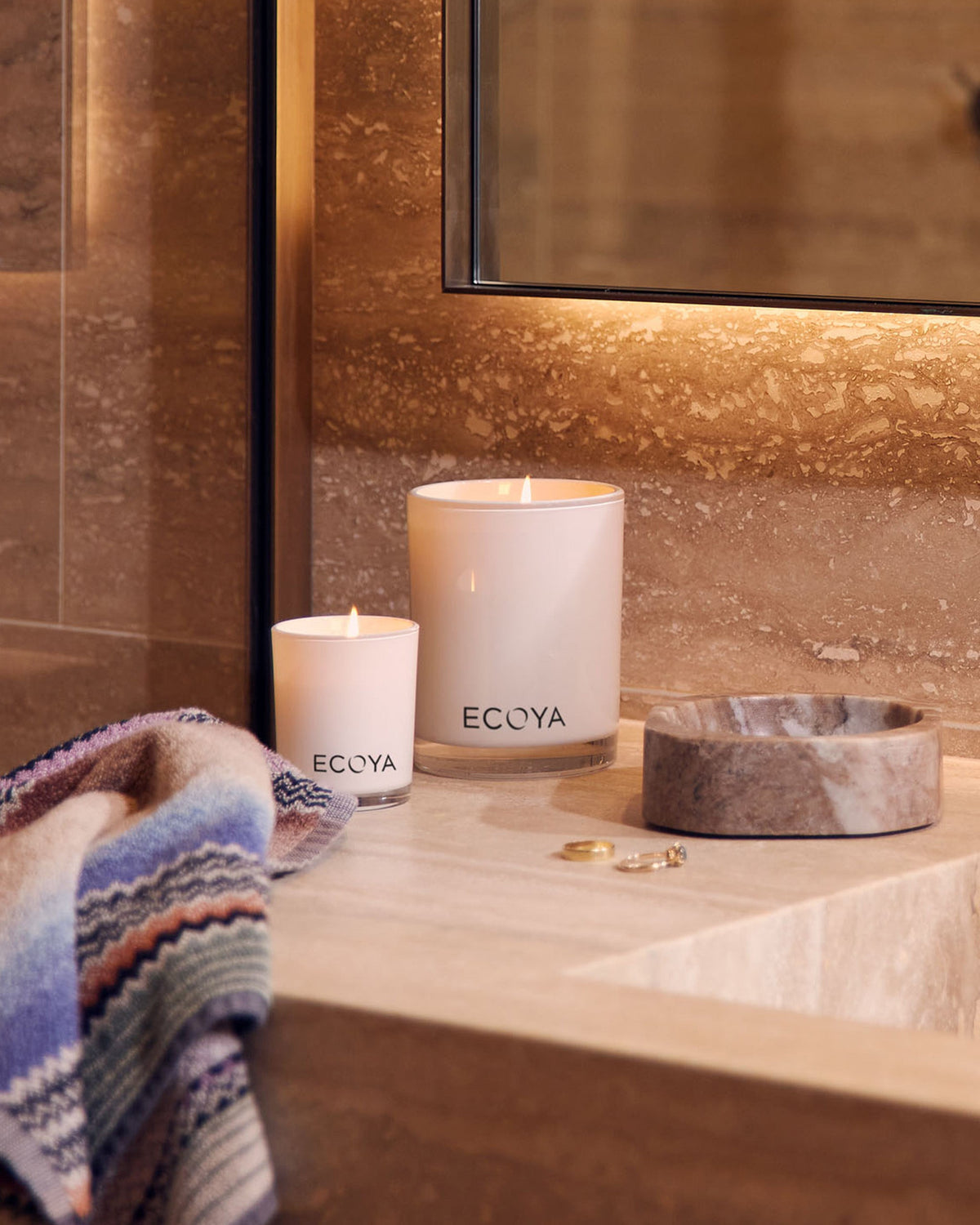 A lit Ecoya Madison candle and a mini candle on a travertine vanity beside a stone dish and a striped towel
