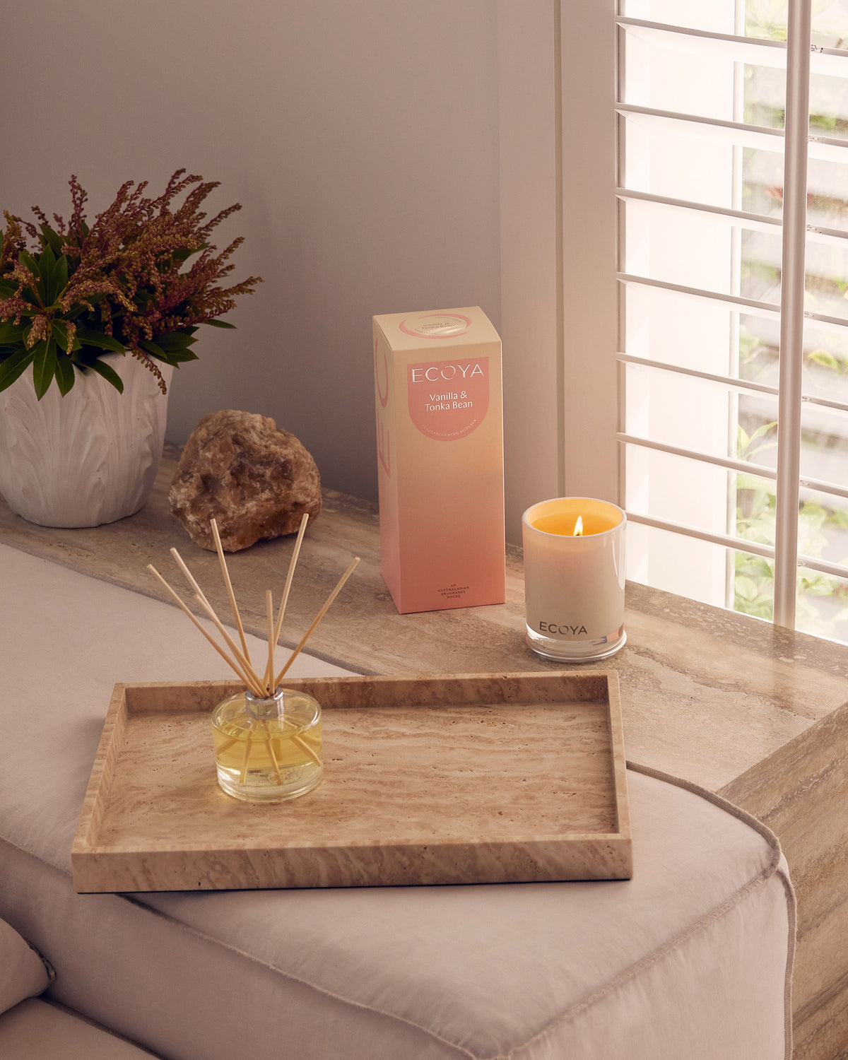 Lit Ecoya candle and matching reed diffuser on a travertine bedside tray