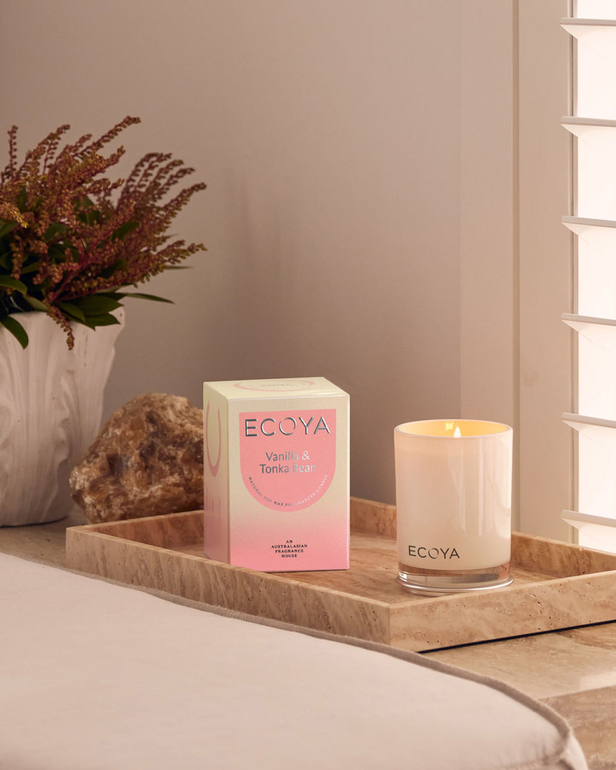 A lit Ecoya Madison candle on a travertine tray with its pink gift box, beside a white vase of dried heather