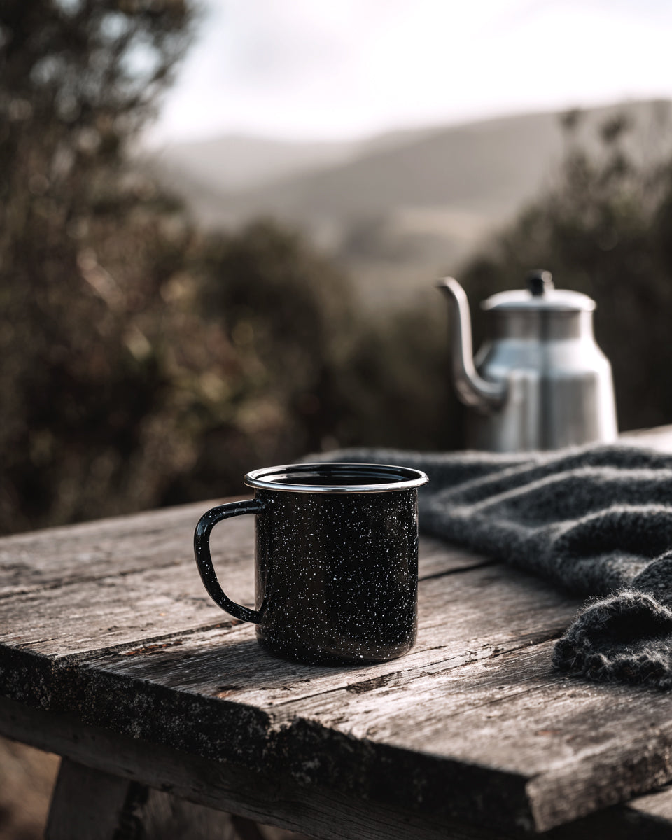 Speckled enamel mug on a weathered outdoor table with a knitted throw and a steel kettle behind