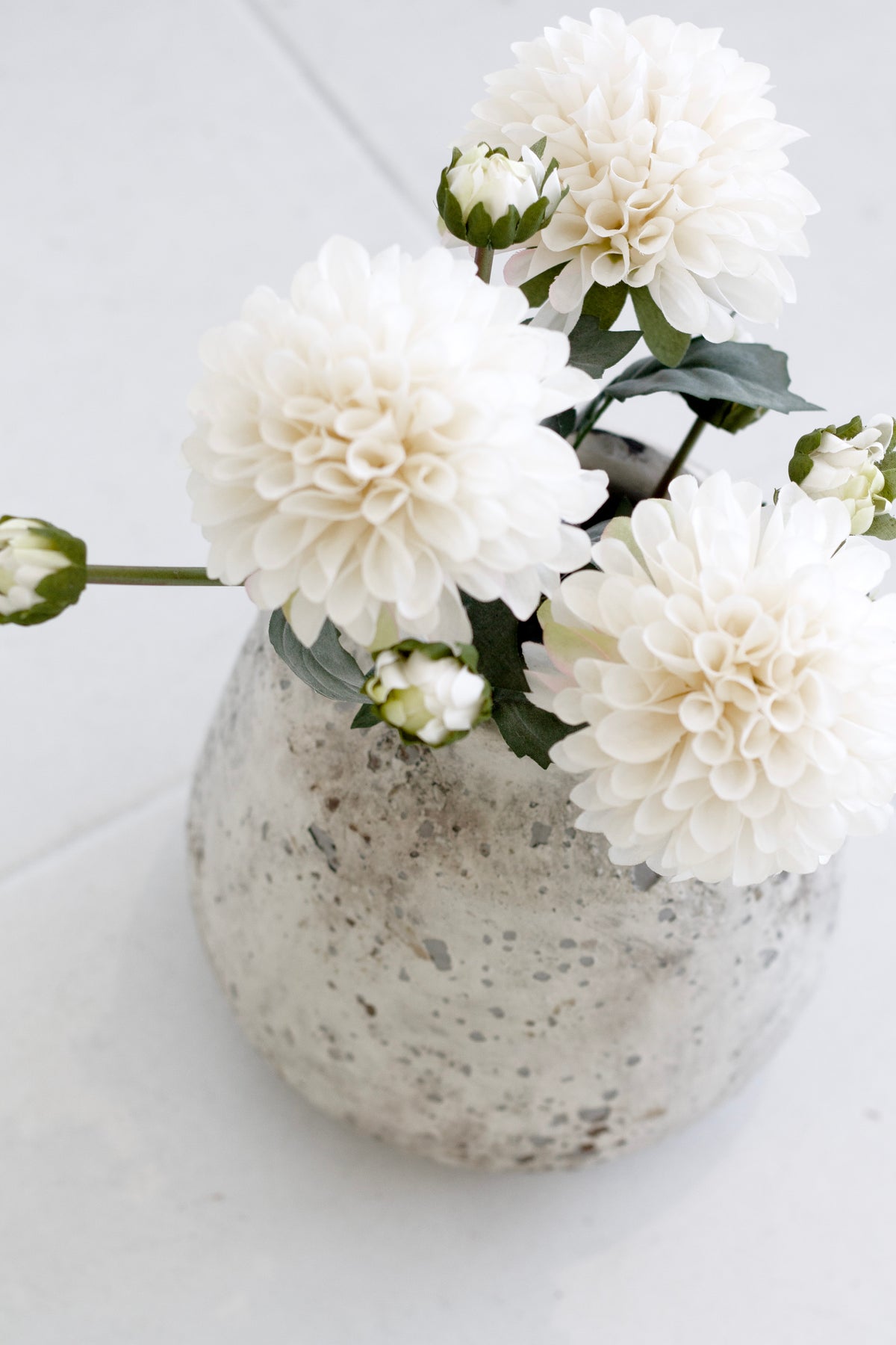 Looking down on dahlias and buds in a speckled stone vase on a pale tiled surface