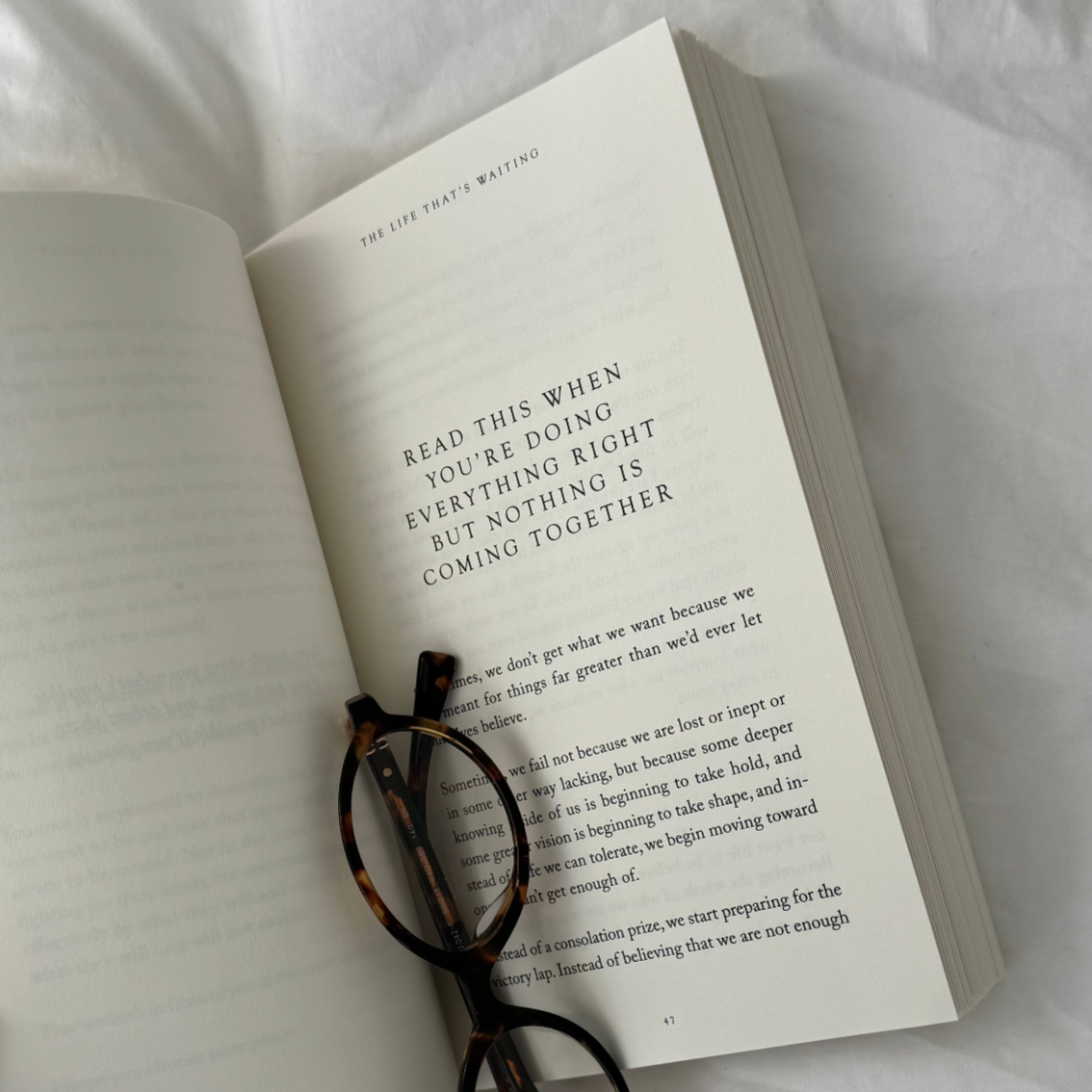 The Life That's Waiting open with tortoiseshell glasses on the page, on white bedding