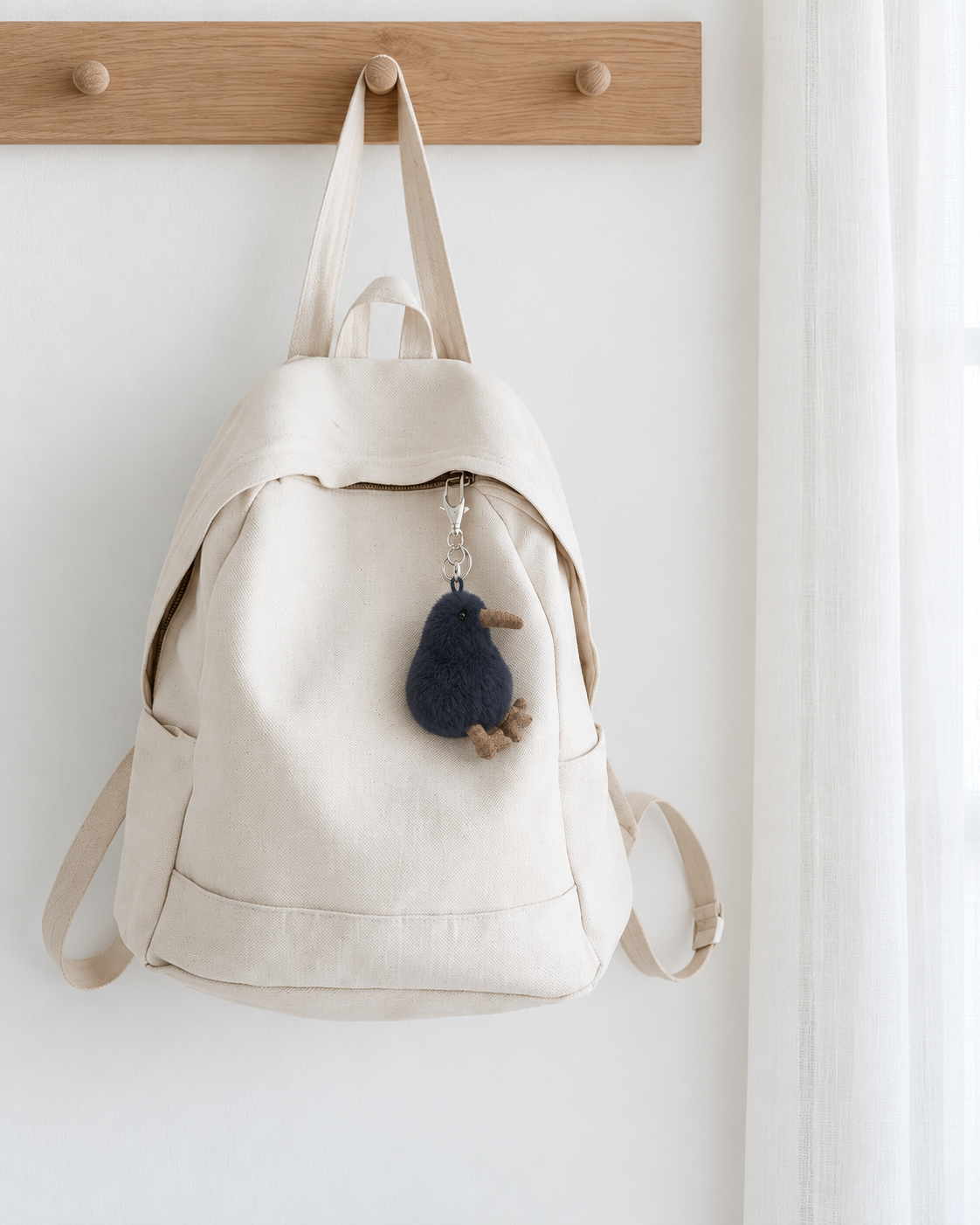 Koko the Kiwi bag charm in Slate by Lily & George clipped to a cream canvas backpack hanging on a timber peg rail