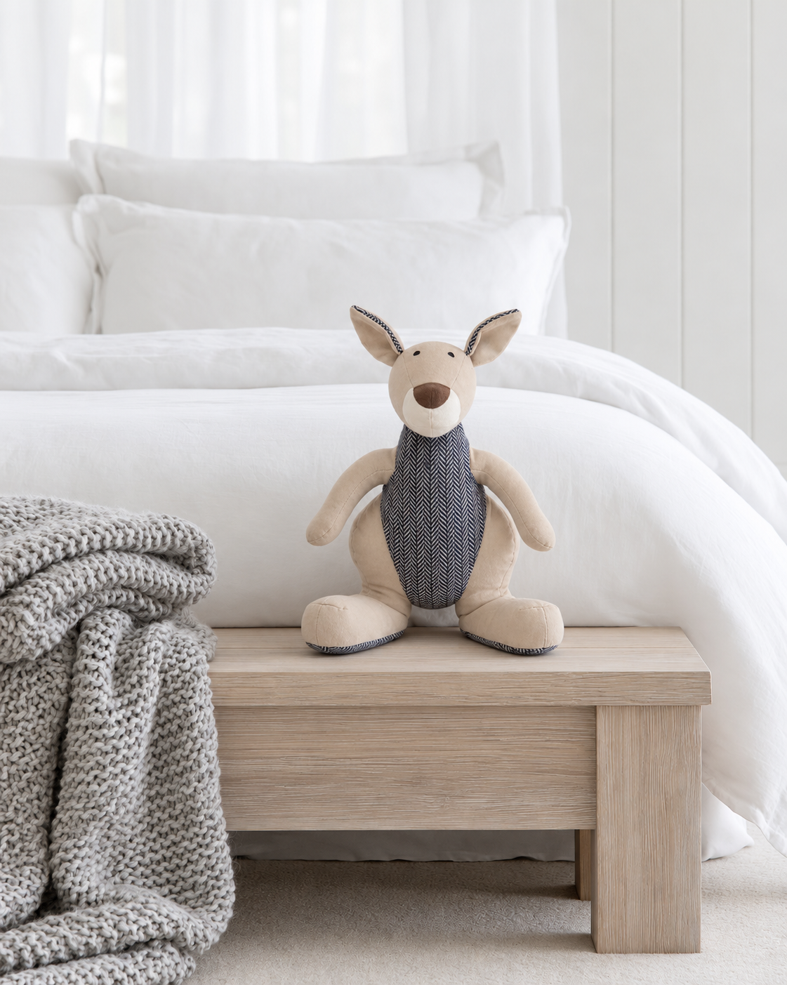 Kazza Kangaroo soft toy by Lily & George standing on a timber bench at the foot of a white bed