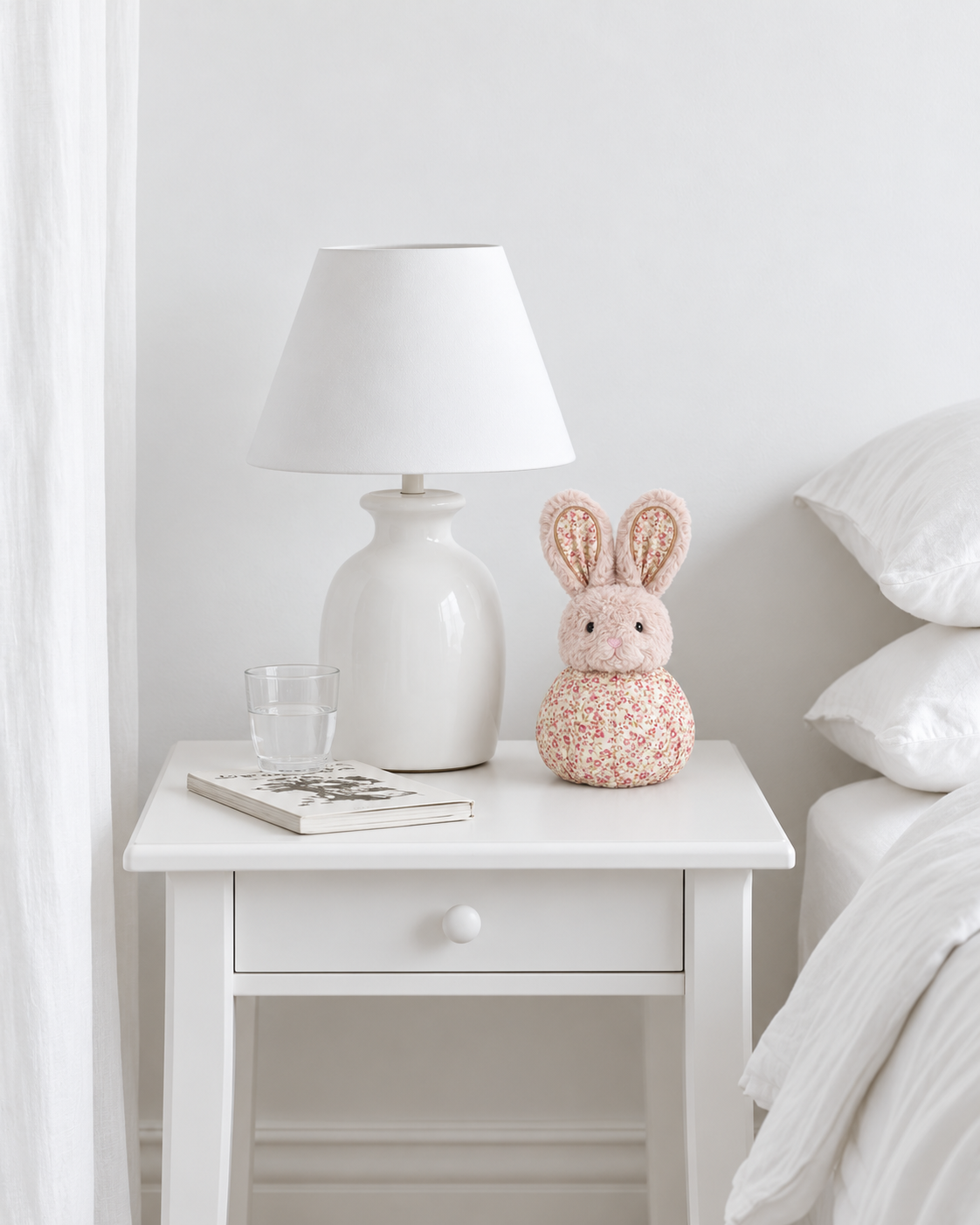 Bailee ball comforter bunny by Lily & George sitting on a white bedside table beside a lamp and a book