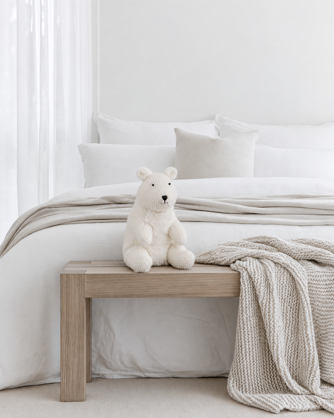 Peter the Polar Bear knitted soft toy by Lily & George sitting on an oak bench at the foot of a white bed