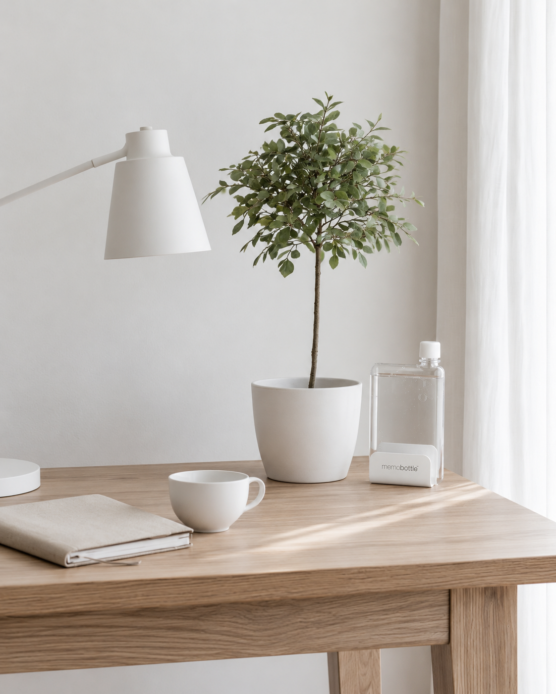 White Memobottle universal desk stand holding a clear flat bottle on an oak desk beside a potted tree